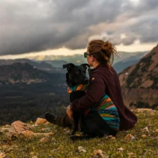 A woman sits on a grassy mountain peak hugging a black dog both looking out over a vast valley of mountains.